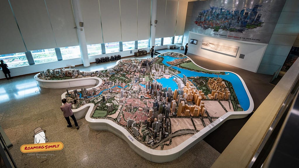 High-angle photograph of the massive 3D architectural scale model of downtown Singapore and the Marina Bay area inside the Singapore City Gallery, with visitors walking around the display.