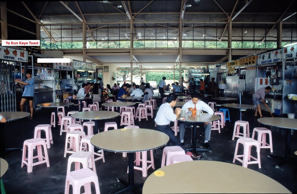 A vintage photograph of the original Ya Kun Kaya Toast stall inside a traditional open-air hawker center in Singapore, with patrons dining at round tables.