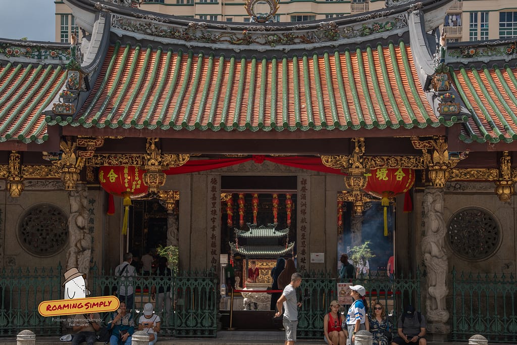 Front entrance of Thian Hock Keng Temple showing the ornate tiled roof, gold-gilded wood carvings, and red lanterns, with visitors gathering at the gate and sitting on the steps.