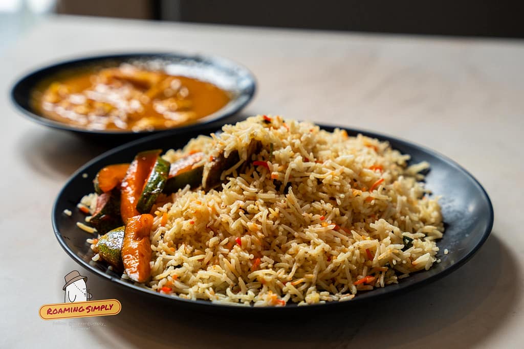 Close-up photograph of a generous portion of Mutton Biryani on a black plate, featuring fragrant long-grain basmati rice and a side of pickled vegetables, with a blurred bowl of curry in the background at Singapore Zam Zam.