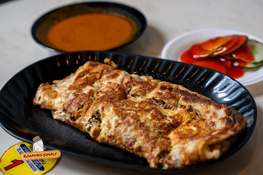 Close-up photograph of a golden-brown, pan-fried Murtabak cut into rectangular slices on a black plate, served with a bowl of curry and cucumber slices at Singapore Zam Zam restaurant.