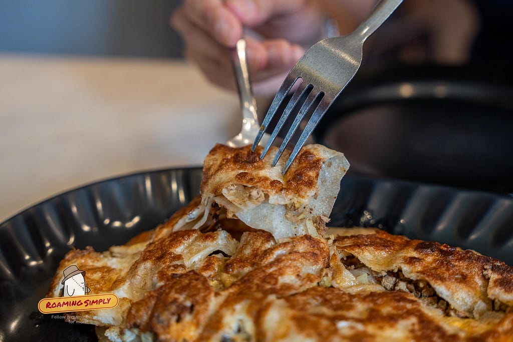 Close-up photograph of a fork lifting a square slice of crispy deer murtabak, revealing the savory minced venison filling inside the golden-brown pan-fried dough on a black plate at Singapore Zam Zam.