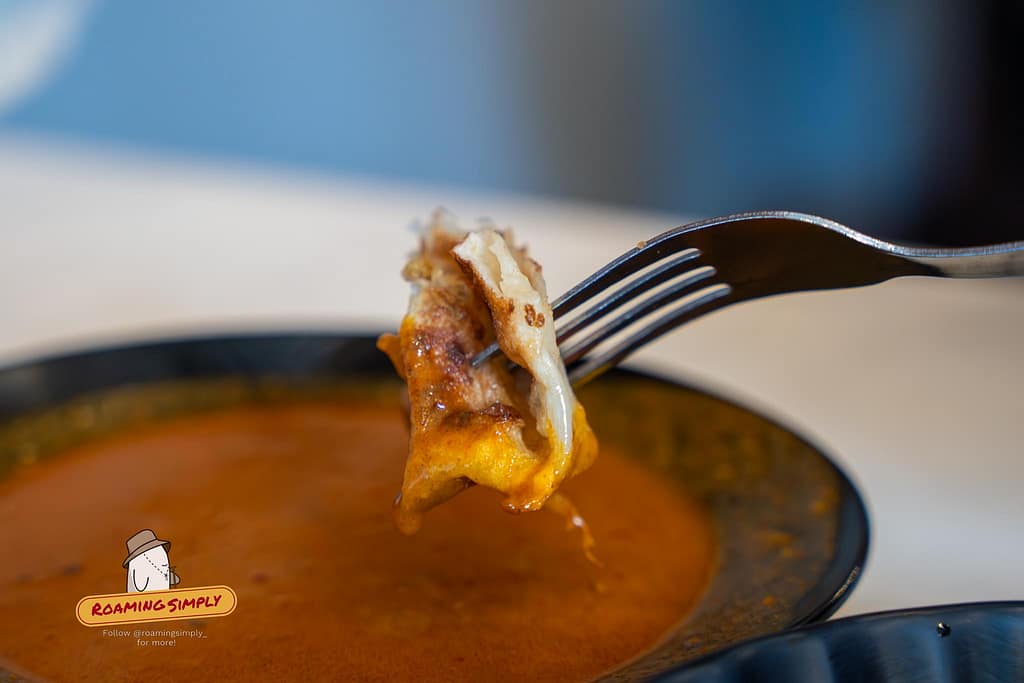 Close-up photograph of a metal fork holding a piece of crispy murtabak dripping with thick, reddish-orange curry sauce over a dark bowl at Singapore Zam Zam.
