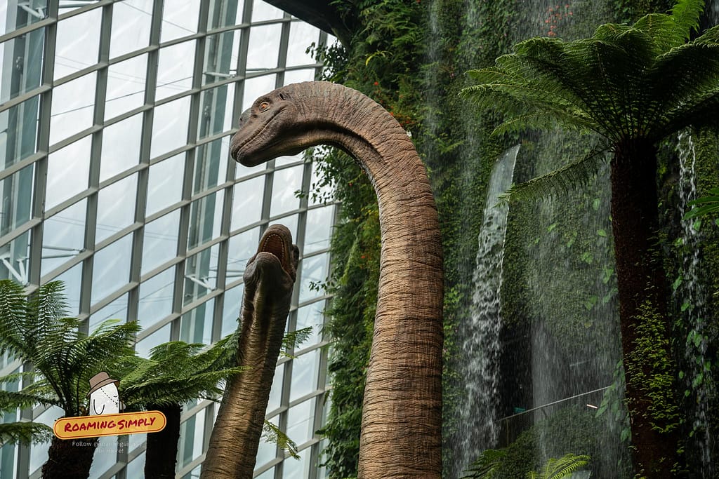 Low-angle shot of two large brachiosaurus dinosaur statues standing in front of a cascading indoor waterfall and lush vertical gardens inside the Cloud Forest glass dome in Singapore.