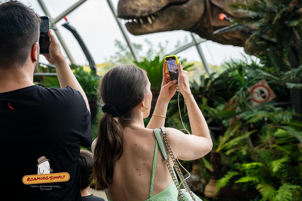 Over-the-shoulder shot of a man and a woman using their smartphones to photograph a massive animatronic T-Rex dinosaur head surrounded by lush greenery inside the Cloud Forest glass dome at Gardens by the Bay, Singapore.