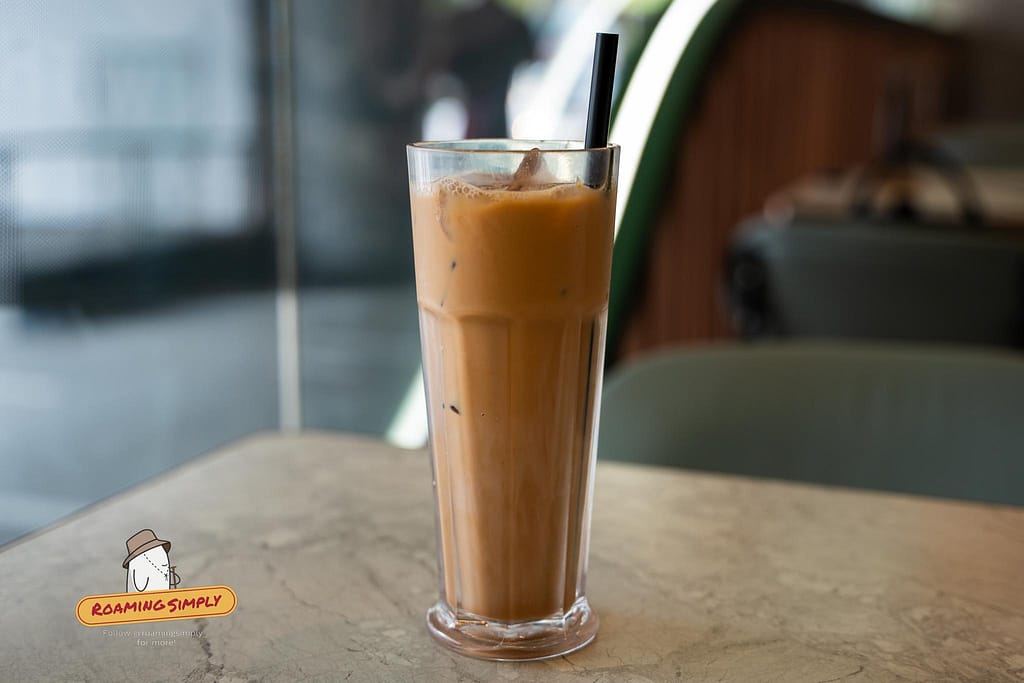 Close-up photograph of a tall glass of iced Hong Kong-style milk tea with condensation and a black straw, resting on a light-colored table at Tsui Wah restaurant against a softly blurred background.