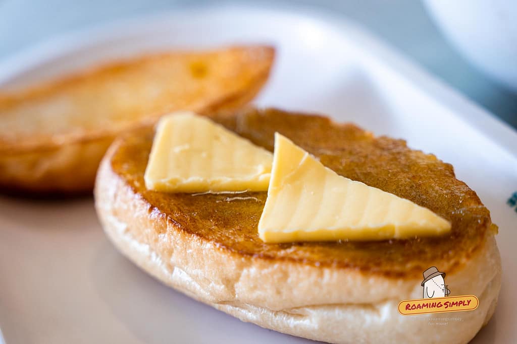 Close-up photograph of a golden-brown toasted bun topped with two thick, triangular slices of butter, glazed with sweet condensed milk and kaya at Tsui Wah restaurant.
