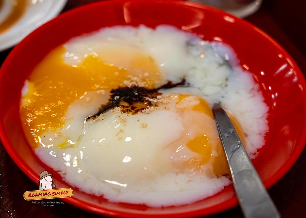 Close-up of Singaporean soft-boiled eggs seasoned with dark soy sauce and white pepper in a red bowl.
