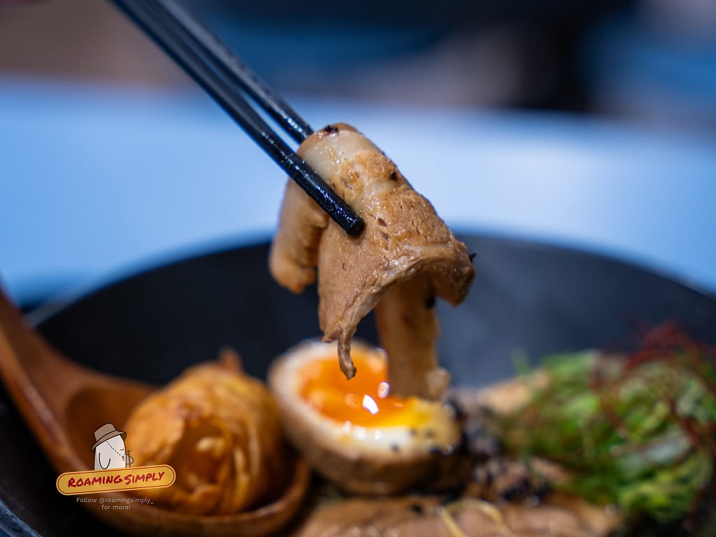 Detailed close-up of tender braised chashu pork belly being held with chopsticks over a bowl of Singapore style ramen, RoamingSimply travel guide and food review.
