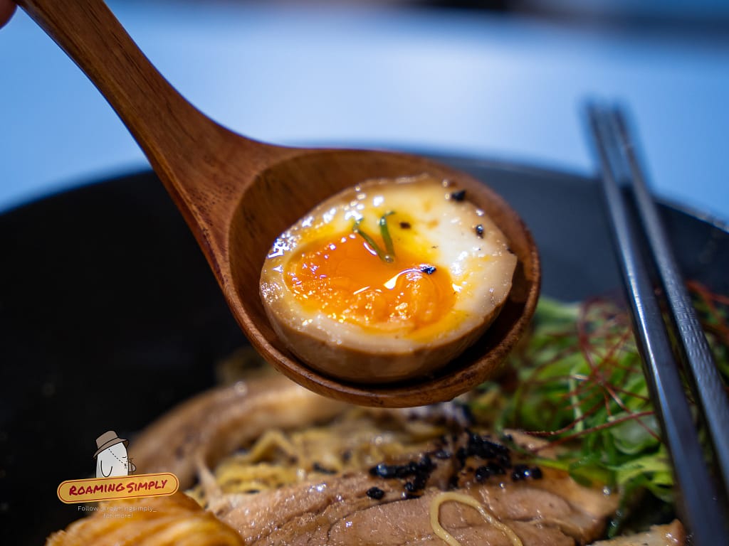 Close-up of a soft-boiled marinated ajitsuke tamago egg on a wooden spoon, served as a creamy topping for Singapore-style ramen, RoamingSimply food review.
