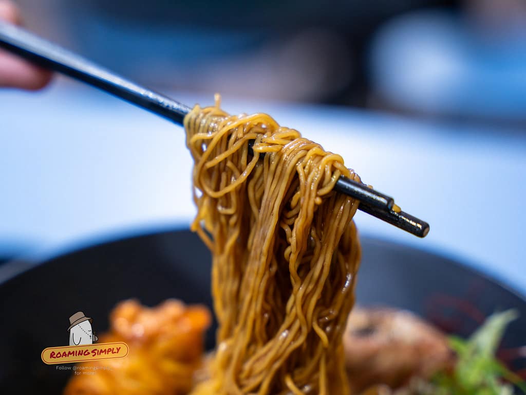 Detailed close-up of thin, springy noodles being lifted with chopsticks from a bowl of Singapore-style ramen, RoamingSimply food review and travel guide.