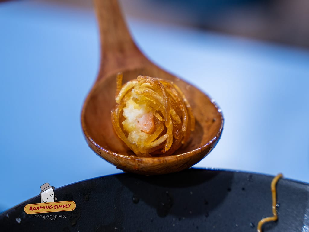 Close-up of a potato-wrapped prawn on a wooden spoon at A Noodle Story Singapore, a unique deep-fried twist on wanton for Singapore-style ramen.
