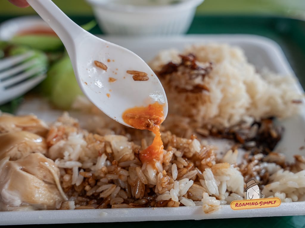 Close-up action shot of a plastic spoon dripping bright orange chili sauce onto a plate of Ah Tai Hainanese Chicken Rice mixed with dark soy sauce.