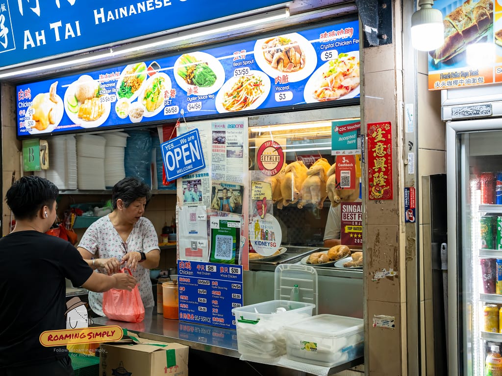 Storefront of Ah Tai Hainanese Chicken Rice at Maxwell Food Centre, featuring a blue signboard with menu photos, hanging poached chickens in the glass display, and a queue of customers.