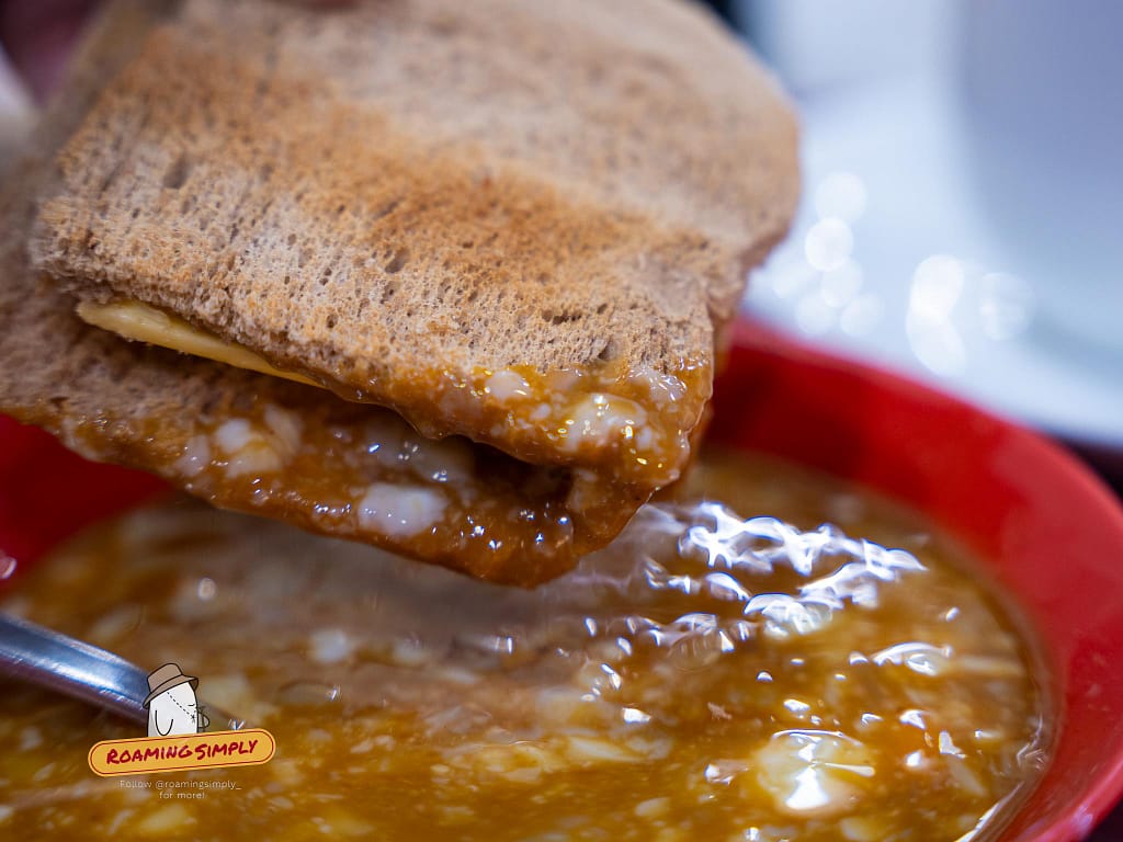 Close-up of crispy Ya Kun Kaya Toast being dunked into a red bowl of seasoned soft-boiled eggs in Singapore.