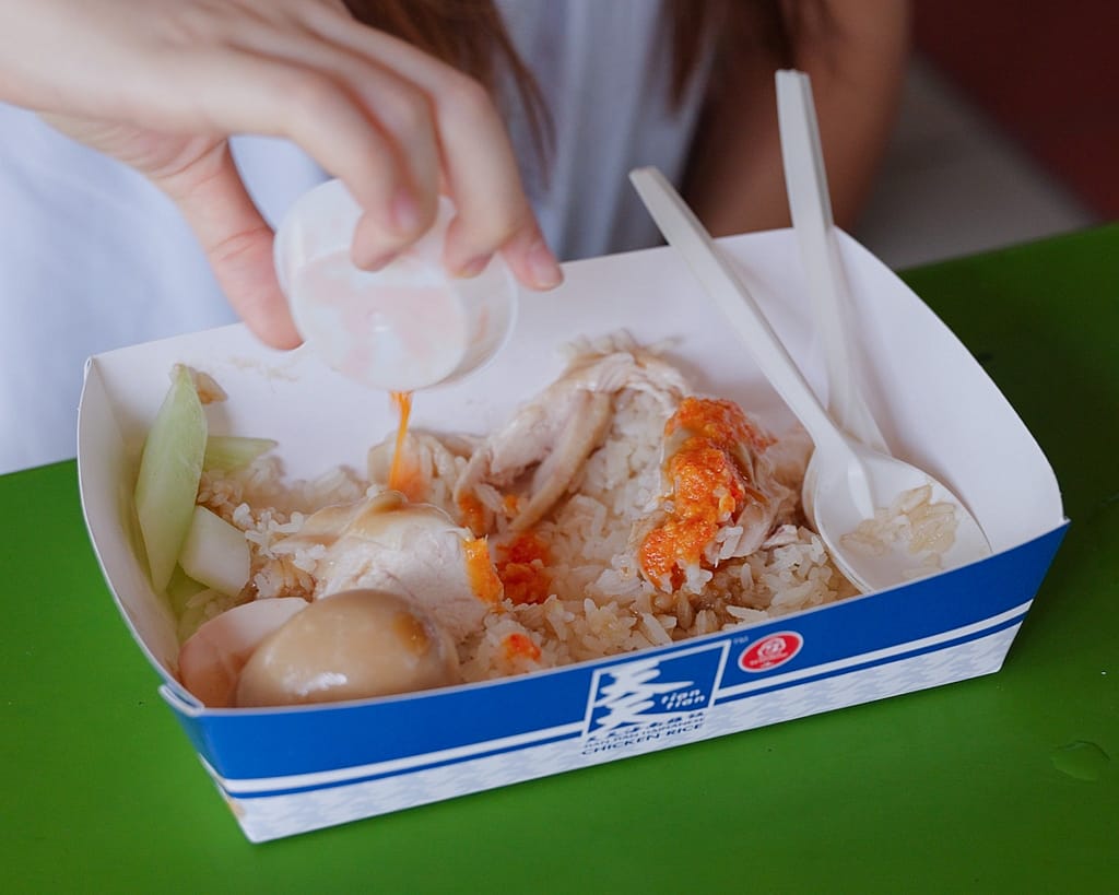 Adding spicy chili sauce to Hainanese chicken rice at a Tian Tian takeaway box in Singapore.