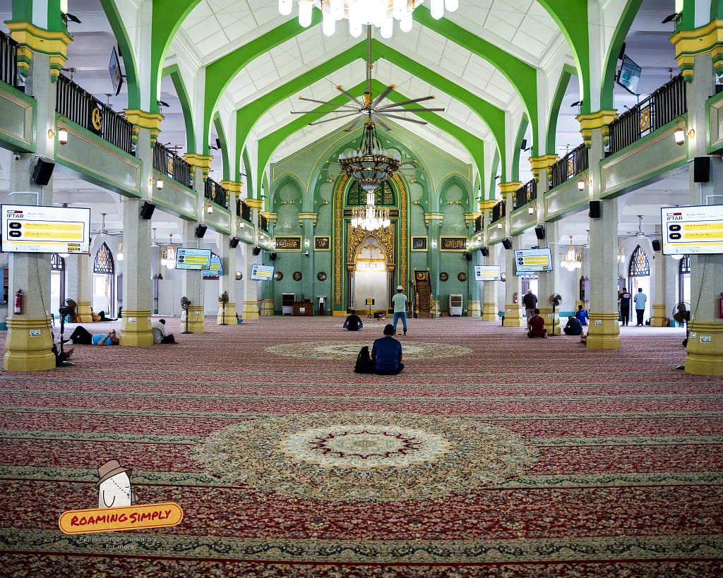 Interior of the grand prayer hall at Sultan Mosque in Singapore, featuring striking green arches, patterned red carpets, and worshippers.