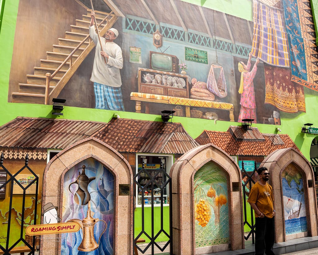 Vibrant heritage street art on a bright green wall in Kampong Glam, Singapore, depicting traditional village life and textile trading above detailed mosaic arches