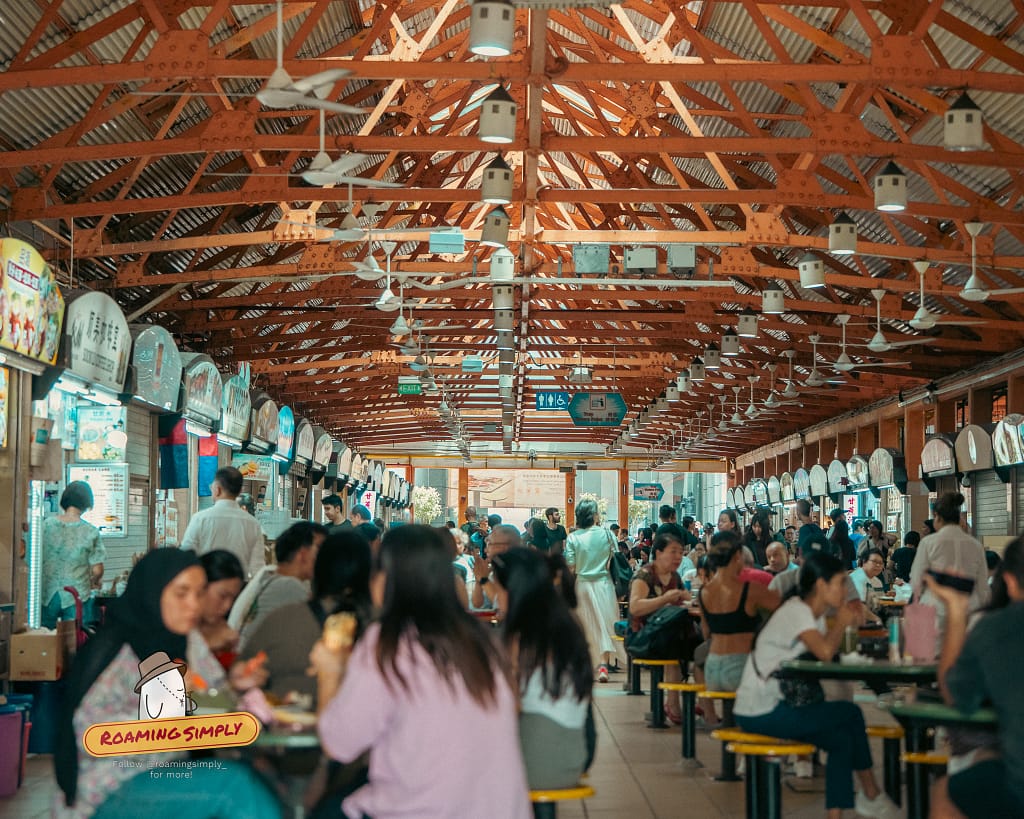 Interior wide shot of Maxwell Food Centre featuring high orange steel roof trusses, ceiling fans, rows of hawker stalls, and a bustling crowd of diners sitting at green round tables.