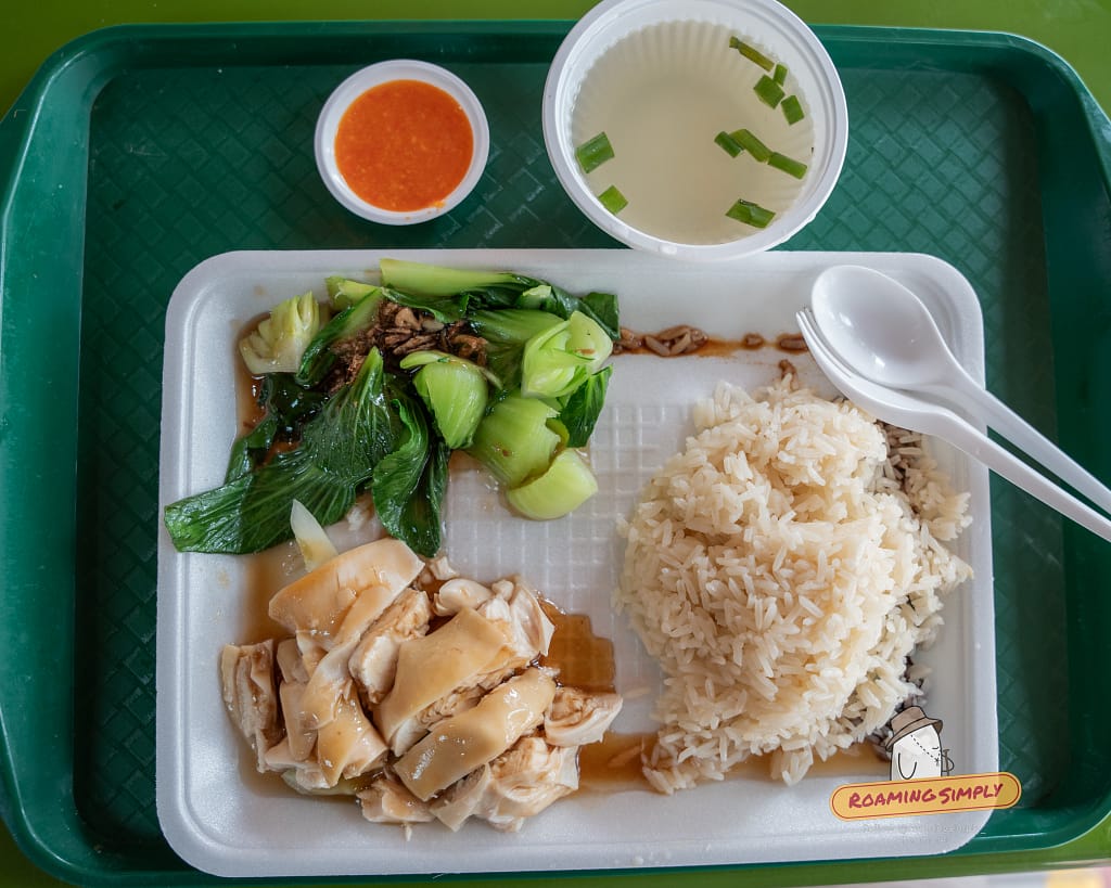 Top-down view of a Hainanese Chicken Rice set meal on a green tray, featuring steamed chicken with soy sauce, rice, blanched vegetables, a bowl of soup, and chili sauce.