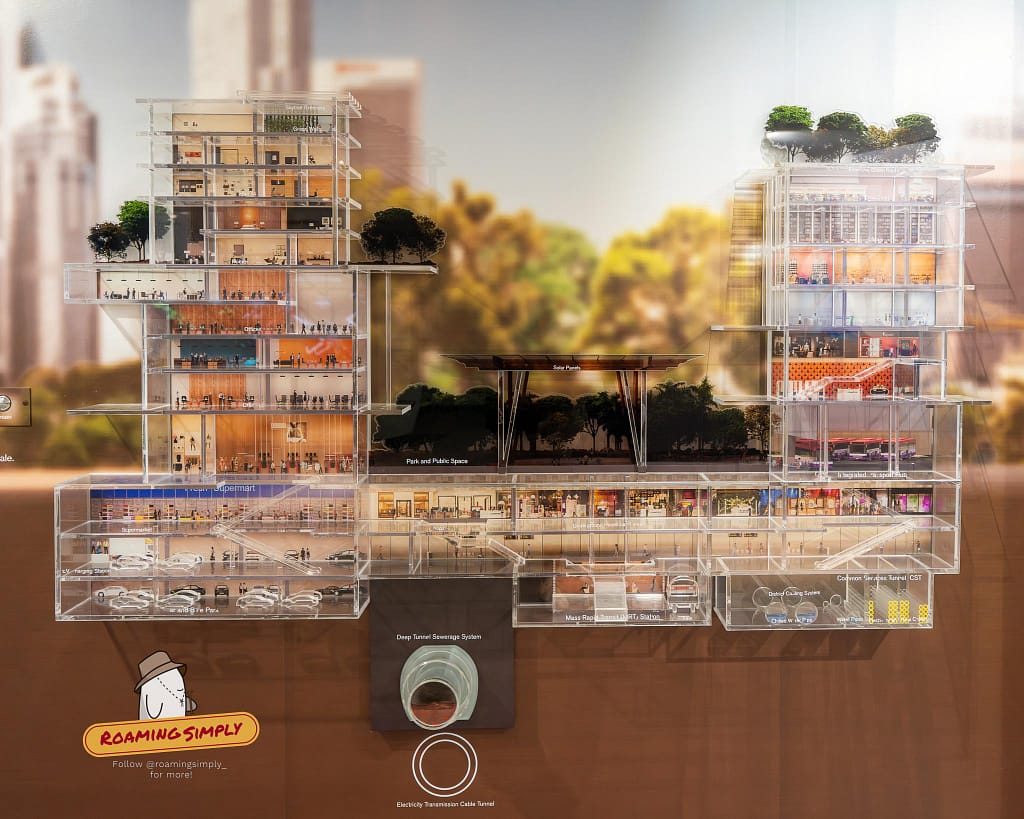 Close-up photograph of a clear acrylic cross-section scale model at the Singapore City Gallery, detailing high-rise buildings with rooftop greenery above deep underground urban infrastructure like utility tunnels and an MRT station.