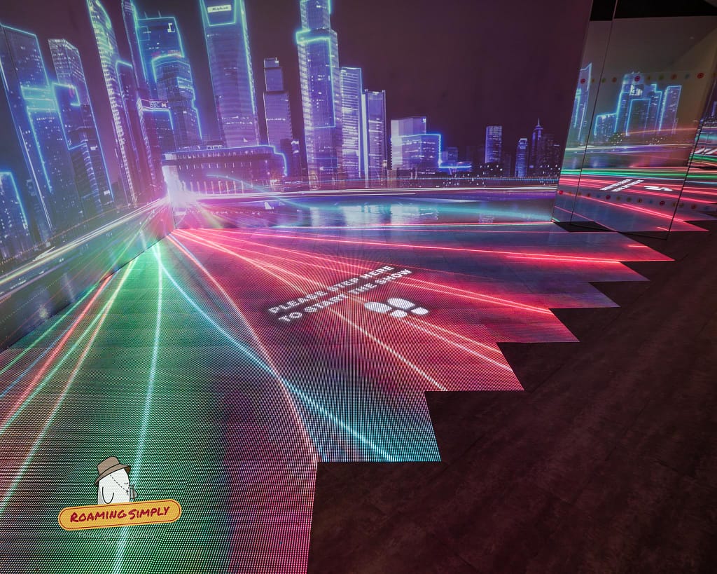 Photograph of an immersive 3D interactive display room at the Singapore City Gallery, featuring neon lights projecting a futuristic city skyline and floor footprints that read 'Please step here to start the show'.
