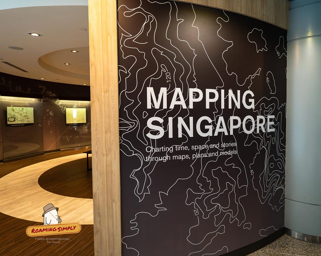 Photograph of the entrance to the Mapping Singapore exhibit at the Singapore City Gallery, featuring a black wall with white topographic lines and text reading 'Mapping Singapore: Charting time, space and stories through maps, plans and models.'