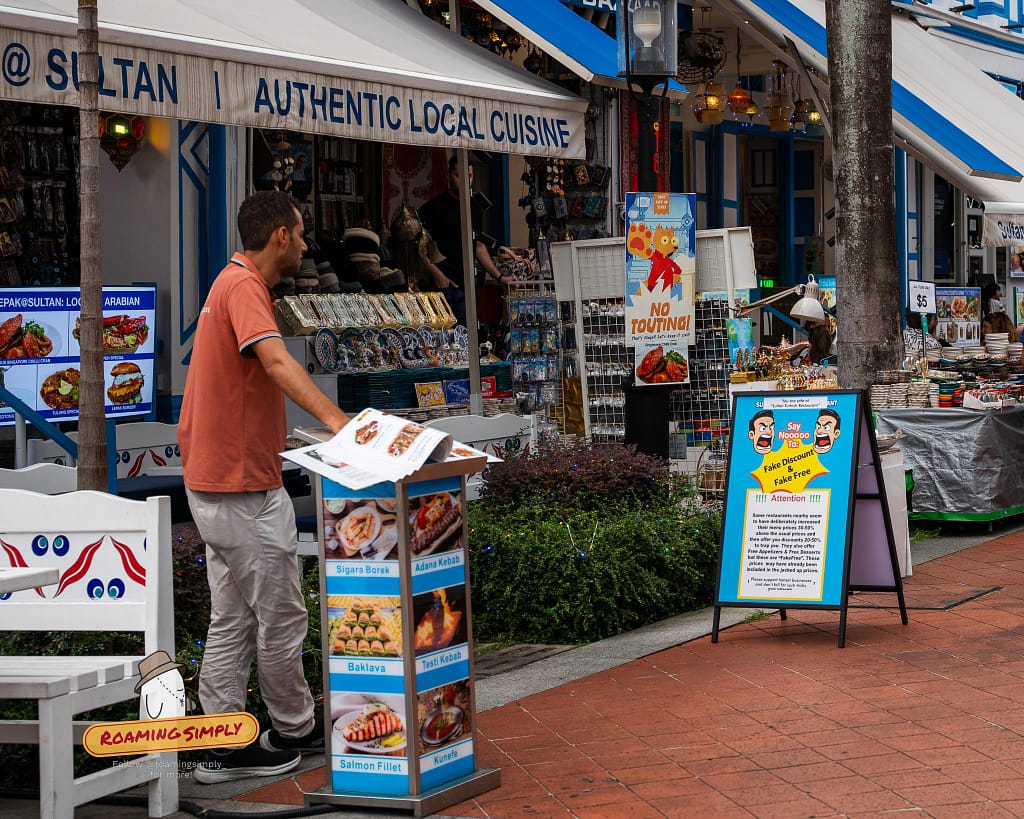 A restaurant host standing by a menu stand on Bussorah Street in Kampong Glam, Singapore, next to prominent signs warning tourists about touting and fake discounts.
