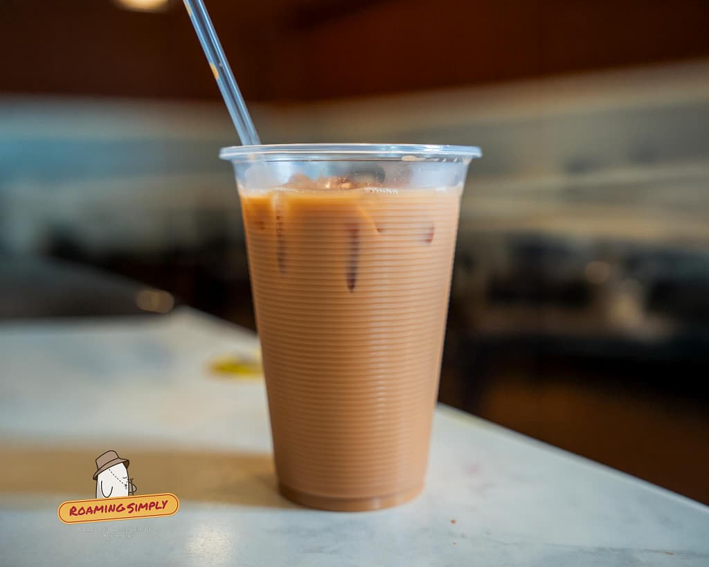 Close-up photograph of a cold, sweet iced Teh Tarik served in a ribbed plastic cup with a straw on a table at Singapore Zam Zam, set against a softly blurred background.