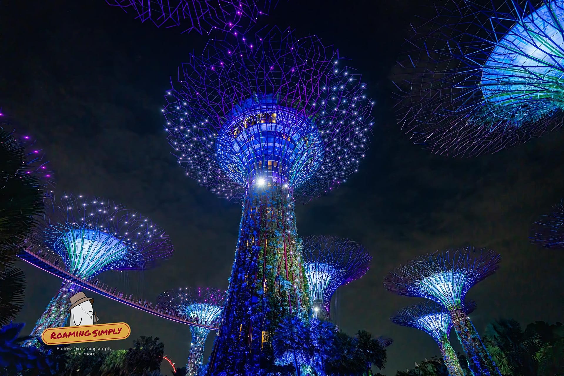 Low-angle night photograph of the towering Supertrees illuminated in vivid blue and purple lights during the Garden Rhapsody show at Gardens by the Bay, Singapore, with the elevated walkway visible.
