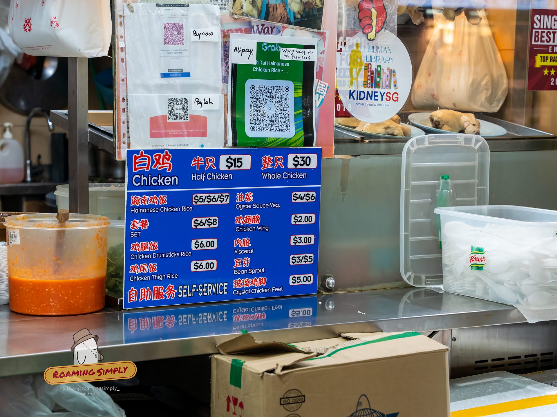 Close-up of the blue menu board at Ah Tai Chicken Rice showing prices for chicken rice sets ($6/$8), whole chicken ($30), and vegetable sides, along with digital payment QR codes.